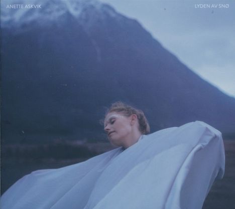 „ANETTE ASKVIK, LYDEN AV SNØ“ oben. Frau in weißem Tuch, Berg im Hintergrund, blauer Farbton.