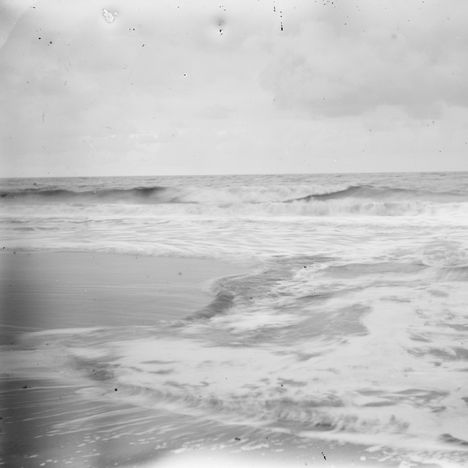 Ein schwarz-weißes Foto zeigt Wellen am Strand unter einem bewölkten Himmel.