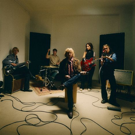 Eine fünfköpfige Bandprobe; Musiker mit Schlagzeug, Bass, Gitarren, Kabel auf dem Boden, lässige Kleidung.