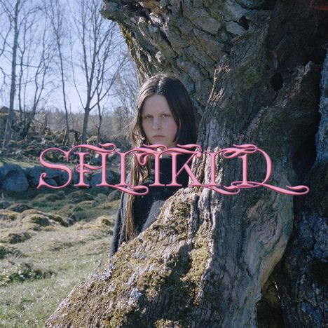 Text: "SMEKKIÐ" in pinker Schrift. Eine Person steht hinter einem Baum in einer Waldlandschaft.