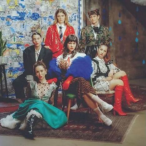 Es gibt keinen erkennbaren Text. Sechs Personen in bunter, modischer Kleidung posieren in einem Raum vor einer abstrakten Wand.