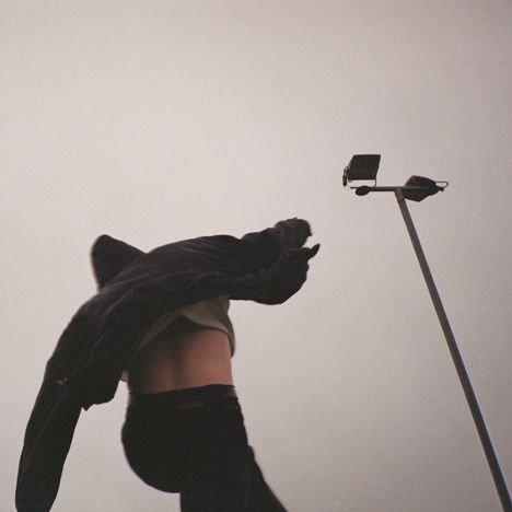 Ein Mensch zieht einen Pullover über den Kopf. Rechts steht ein hoher Lichtmast. Der Himmel ist grau und wolkenverhangen.