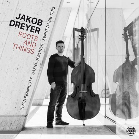 Text: "JAKOB DREYER ROOTS AND THINGS" und Namen von Musikern. Ein Mann steht mit einem Kontrabass in einem modernen Flur.