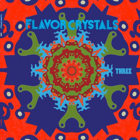 "FLAVOR CRYSTALS" und "THREE" in Blau. Bunte, symmetrische Mandala-Illustration mit kräftigen Farben und Mustern.