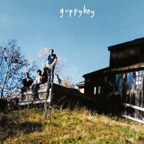 Text: "guppyboy". Eine Gruppe von Personen sitzt und steht auf einer Terrasse vor einem hölzernen Gebäude in ländlicher Umgebung.