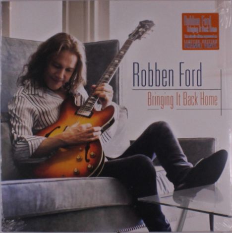 "Robben Ford Bringing It Back Home." Ein Mann mit langer Haaren spielt eine Gitarre auf einem Sofa.