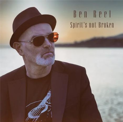 Ben Reel, "Spirit's not Broken". Ein Mann mit Brille und Hut am Wasser, trägt ein T-Shirt mit Gitarrenmotiv.