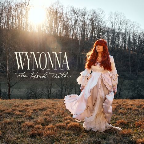 Text: "WYNONNA The Hard Truth". Eine Frau mit rotem Haar in einem langen weißen Kleid steht auf einem Feld bei Sonnenuntergang.