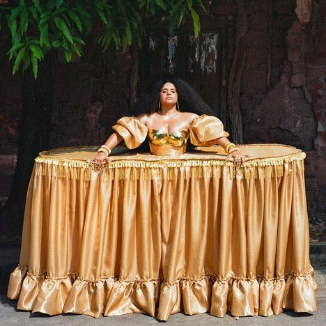 Ich kann den Text im Bild nicht erkennen. Eine Frau trägt ein goldenes, tischähnliches Kleid mit voluminösen Ärmeln.