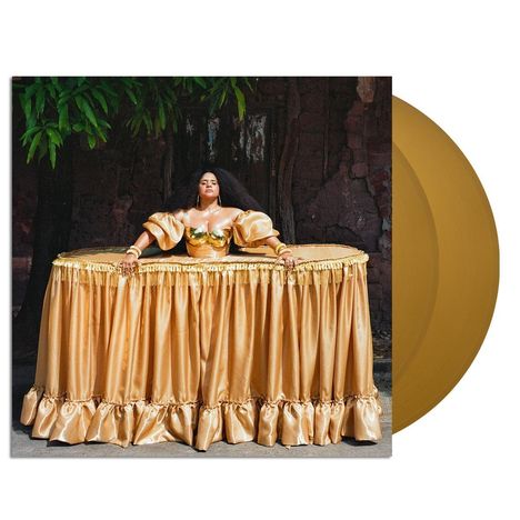 Eine Person in einem goldenen, ausladenden Kleid steht vor einer dunklen Wand. Daneben eine goldene Schallplatte.