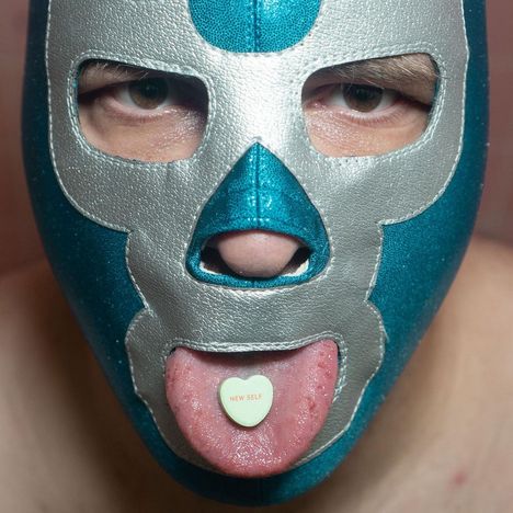 Herz mit "NEW SELF" auf Zunge, Person trägt türkis-silberne Wrestling-Maske und schaut direkt nach vorne.