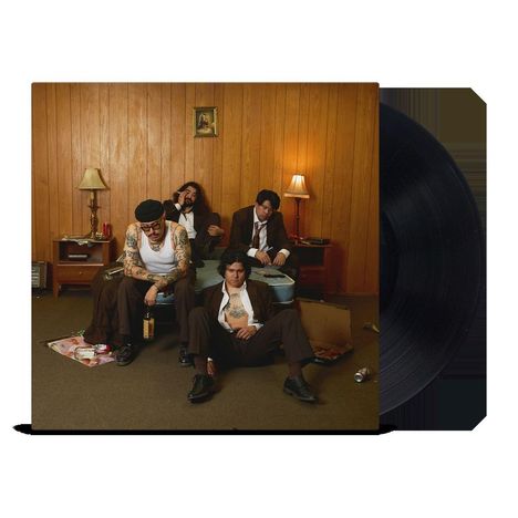 Vier Männer in Anzügen in einem rustikalen Wohnzimmer mit Holzwänden und Lampen, ein kleiner Bilderrahmen an der Wand.