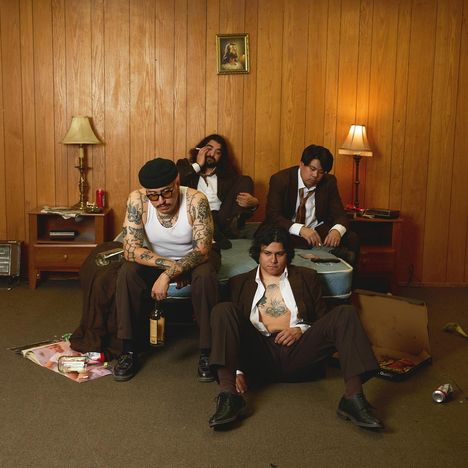 Vier tätowierte Männer in einem holzgetäfelten Raum sitzen entspannt; Chaos und leere Flaschen umgeben sie.