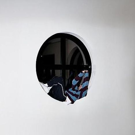 Ein rundes Fenster mit schwarzem Rahmen. Dahinter hängen zwei gestreifte Pullover in dunkelblauen und violetten Tönen.