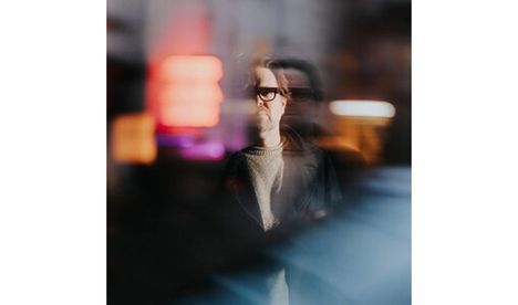 Ein unscharfes Farbfoto zeigt das verschwommene Gesicht eines Mannes mit Brille hinter einer Scheibe. Neonlichter im Hintergrund.