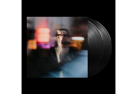 Unschärfe eines Mannes mit Brille, hellem Pullover, Neonlichter spiegeln sich. Dahinter schwarze Schallplatten.