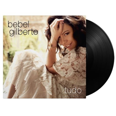 Text: "bebel gilberto" und "tudo". Eine Frau in einem Spitzenkleid sitzt lächelnd, eine Hand am Kopf. Vinyl-Schallplatte sichtbar.