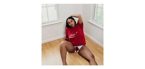 Der Text auf dem roten T-Shirt lautet "Girls". Eine Frau sitzt auf hellem Holzboden, trägt ein rotes T-Shirt und bedruckte Unterwäsche.