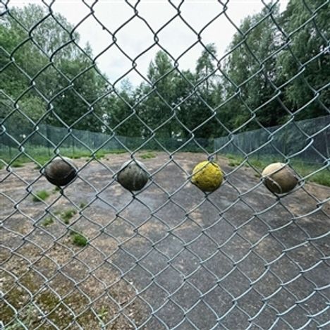 Vier Bälle klemmen in einem Maschendrahtzaun, dahinter ein leerer, überwucherter Tennisplatz.