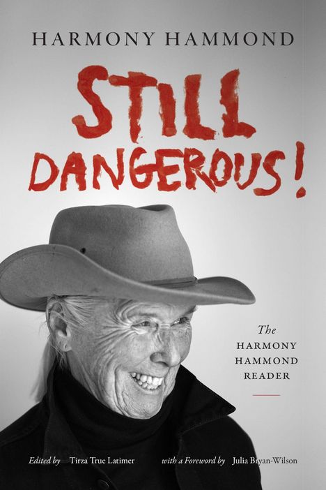 Text: "Harmony Hammond: Still Dangerous! The Harmony Hammond Reader. Edited by Tirza True Latimer with a Foreword by Julia Bryan-Wilson." Schwarz-weiß Porträt einer lächelnden Person mit Hut.