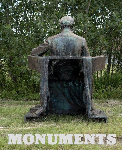 Text: „MONUMENTS“. Eine verwitterte Statue eines sitzenden Menschen von hinten, in einer grünen Umgebung.