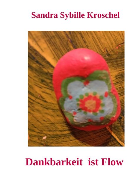 Text: "Sandra Sybille Kroschel", "Dankbarkeit ist Flow". Ein bemalter Stein mit Blumenmotiv auf Holzhintergrund.