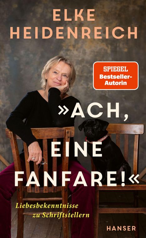 Der Text lautet: "Elke Heidenreich. »Ach, eine Fanfare!« Liebesbekenntnisse zu Schriftstellern. Spiegel Bestseller-Autorin". Eine Frau sitzt lächelnd mit einem schwarzen Hund auf Holzstühlen.