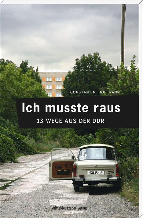 „Ich musste raus: 13 Wege aus der DDR.“ Ein altes Auto steht auf einem verlassenen Weg vor einem Plattenbau.