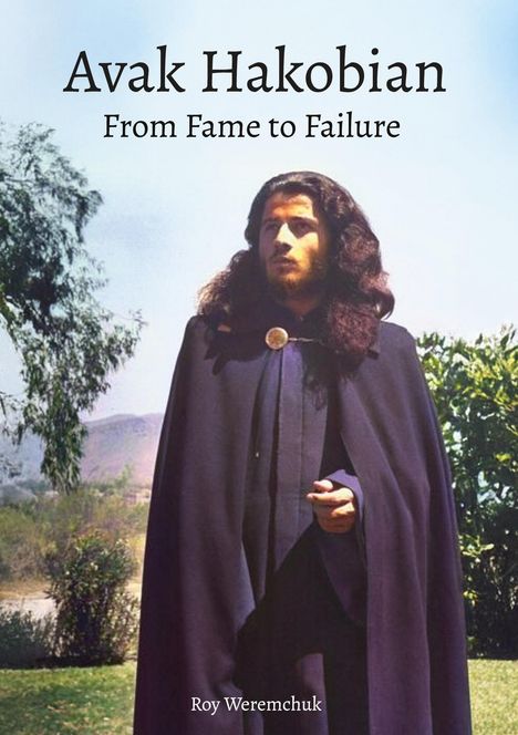 Ein Mann mit langen Haaren und Umhang steht vor einer natürlichen Kulisse mit Bäumen und Bergen.