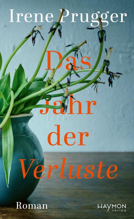 „Irene Prugger, Das Jahr der Verluste, Roman“. Vase mit welken Blumen auf Holztisch vor grauer Wand.