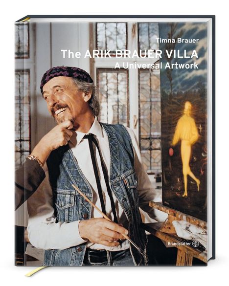"Timna Brauer, The ARIK BRAUER VILLA, A Universal Artwork." Ein Mann in Jeansweste lächelt, hält Pinsel vor Gemälde.