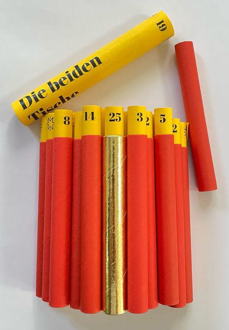 Zylinder mit rotem Papier umwickelt, oben gelbe Enden mit Zahlen. Über einem liegt ein ebenfalls gelber Zylinder mit "Die beiden" und "Tische".