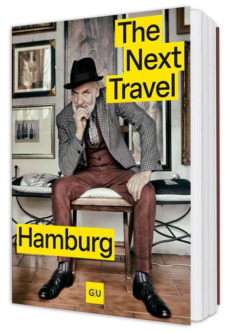 "GU, The Next Travel Hamburg." Ein älterer Mann in Anzug und Hut sitzt auf einem Stuhl vor gerahmten Bildern.