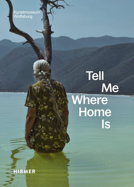 Texte: "Kunstmuseum Wolfsburg", "Tell Me Where Home Is". Eine Frau steht im Wasser, im Hintergrund Berge und ein karger Baum.