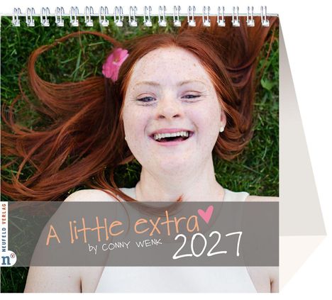 „A little extra by Conny Wenk 2027.“ Eine lächelnde Person mit roten Haaren liegt im Gras, eine rosa Blume im Haar.