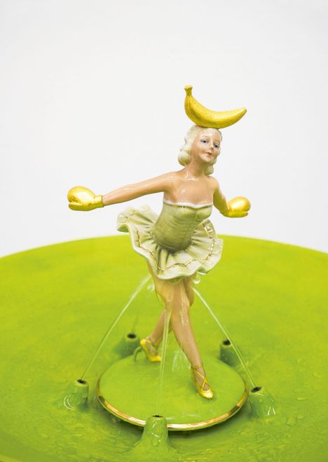 Eine Tänzerin in einem grünen Kleid balanciert eine Banane auf dem Kopf, mit ausgebreiteten Armen.