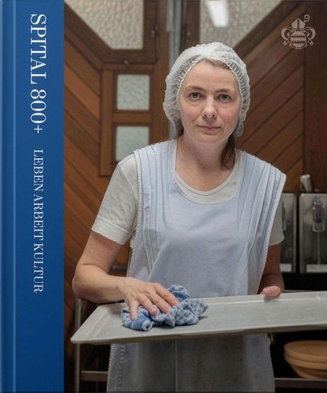 "SPITAL 800+ LEBEN ARBEIT KULTUR". Eine Frau mit Haarnetz und Schürze hält ein Tablett mit einem Tuch in einem Küchenraum.