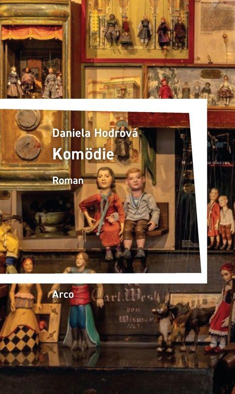 Buchtitel: "Komödie" von Daniela Hodrová. Marionetten in traditioneller Kleidung auf einer Bühne. Darunter steht "Arco".