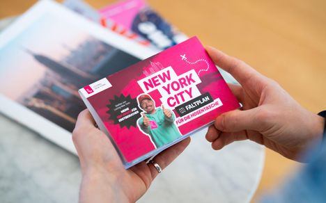 Text: "New York City Faltplan für die Hosentasche". Hände halten den Plan. Im Hintergrund verschwommene Bilder.