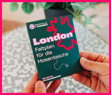„London Faltplan für die Hosentasche.“ Hände halten eine faltbare Karte. Hintergrund mit Pflanzen und Tischmuster.