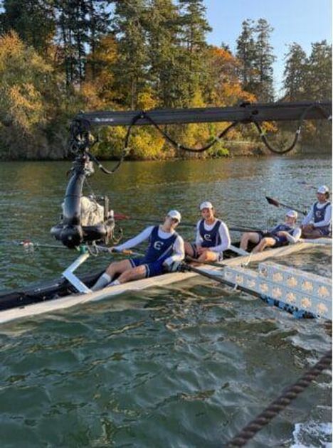 Ruderer in einem Boot auf einem Fluss, umgeben von Bäumen im Herbst. Ein Kamerakran ragt über das Boot.