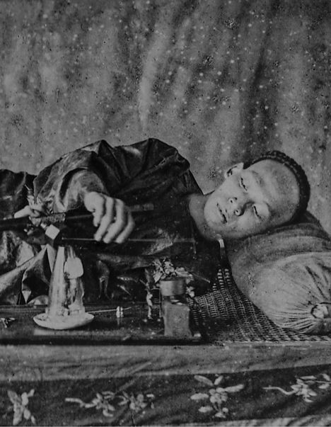 Ein Mann liegt seitlich, stützt sich auf Ellbogen, mit Pfeife in der Hand, traditionelle Kleidung, unscharfer Hintergrund.