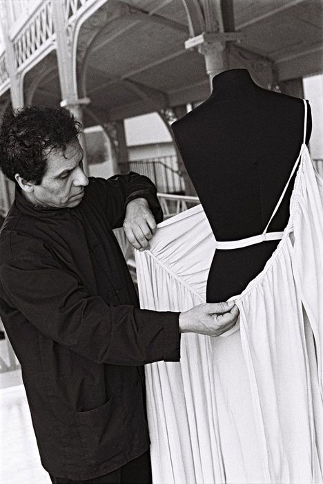 Keine sichtbaren Texte. Ein Mann passt ein langes, helles Kleid an einer schwarzen Schneiderpuppe an. Elegantes Ambiente.
