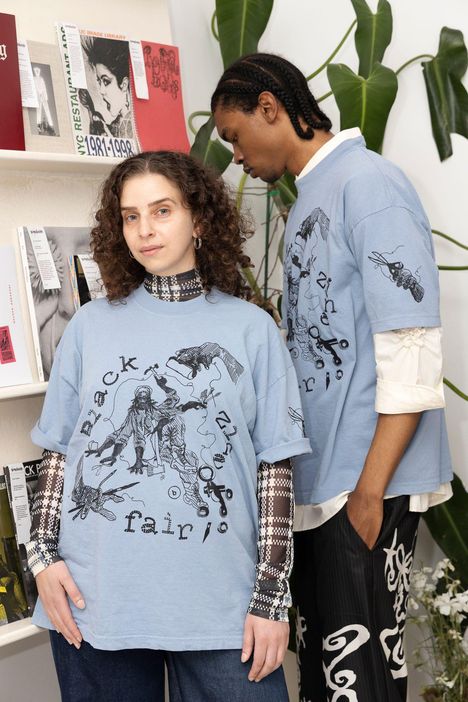 Zwei Personen in blauen T-Shirts mit dem Text "Black fair". Im Hintergrund Regale mit Büchern und Pflanzen.