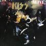 Texte: "KISS", "ALIVE!". Rockband in auffälligen Kostümen auf einer rauchigen, beleuchteten Bühne.