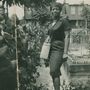 Keine sichtbaren Texte. Eine Frau in einem Garten, trägt ein gestreiftes Kleid und eine Handtasche, lächelt.
