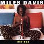 "MILES DAVIS" oben, "doo-bop" unten. Ein Mann auf einem Bett spielt Trompete, umgeben von bunten Kissen und Kunstwerken.