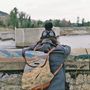 Text auf dem Rucksack: Ein Pfad durch Landschaft. Person mit Wollmütze blickt auf einen Fluss hinter einer Steinmauer.