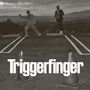 Text "Triggerfinger". Zwei Männer in Anzügen stehen auf einem Parkplatz, einer zeigt mit dem Finger, im Hintergrund Berge.