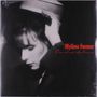 Auf einer Aufnahme steht in roter Schrift "Mylène Farmer, Cendres de lune". Sie zeigt eine Frau in Schwarz-Weiß, die einen Hut anfasst.